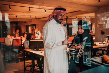 Portrait photo of Arab leader businessman with muslim hijab woman discussing business projects while using tablets and smartphone in modern glass office 