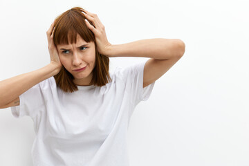 Naklejka premium a funny, emotional woman stands on a light background in a T-shirt and holds her head with her hands while expressing emotions. Horizontal photo with an empty space for inserting an advertising layout