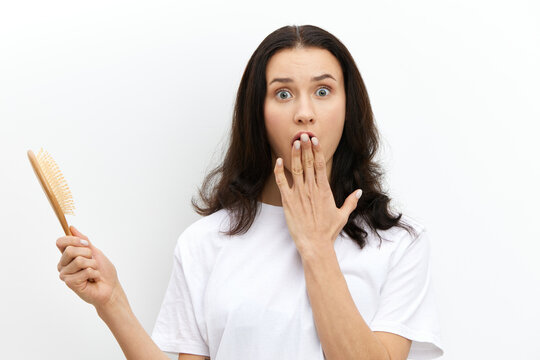 a funny, frightened, emotional woman holds a comb in her hand with her eyes wide open with fright and covers her mouth with her hand. Horizontal photo on a light background with an empty space