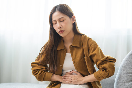 Flatulence asian young woman hand in stomach ache, suffer from food poisoning, abdominal pain and colon problem, gastritis or diarrhoea sitting on sofa at home. Patient belly, abdomen or inflammation.