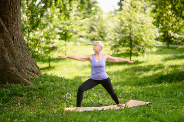 Middle aged woman doing yoga outdoors in warrior pose, Virabhadrasana II. Sports concept in city park.