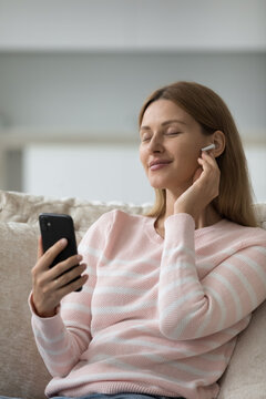 Serene woman holds smartphone listens music through modern earphones, relaxing seated on sofa, enjoy favourite track looks peaceful, spend weekend at home use digital stream services and modern tech