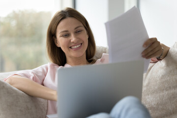 Young millennial 35s woman sits on sofa with laptop, holds receipts or bills, makes payment through e-bank, working at home, doing paperwork, reviewing savings and bank statement smile feel satisfied