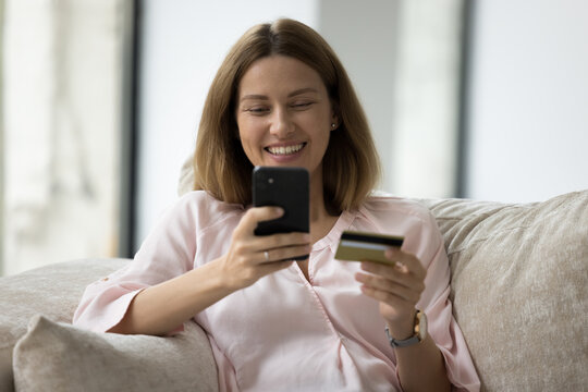 Young smiling woman use cellphone and credit card makes easy secure payments on-line via e-bank app, retail client buy goods on internet, spend received bonus cashback feels happy. Electronic shopping