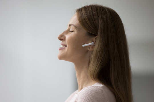 Serene woman listens music through wireless earphones, closeup side face view. Silent tranquil attractive female wear white earbuds enjoy favourite track relish quality of sound, Modern tech, hobby