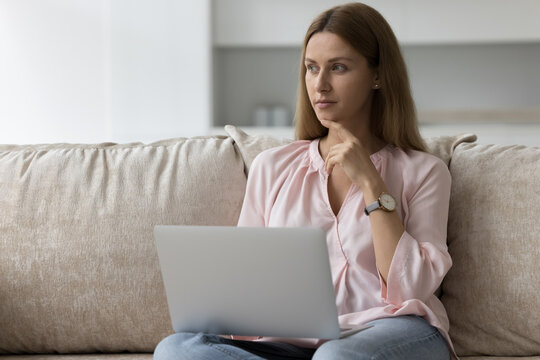 Young woman ponder answer to client working at home using laptop on weekend. 35s female choose goods and services buying on internet sit on sofa with computer looks undecided. Leisure, telework, tech