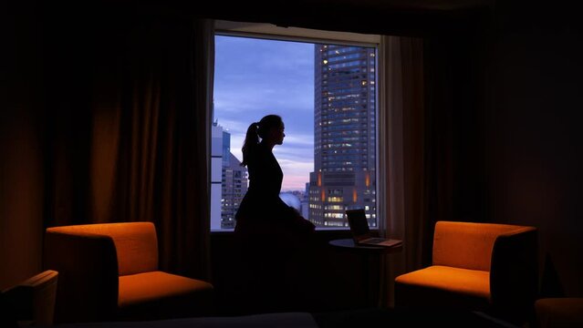 Young woman sitting on window sill in dark room, looking out at the evening city. Blurred background, dark silhouette of pensive lady against the sunset sky. Notebook computer on small table near her