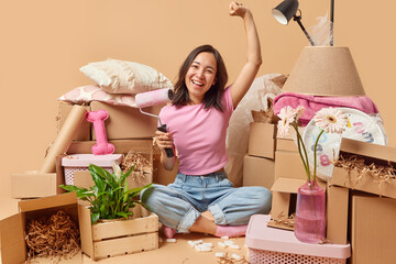 Happy Asian woman going to paint walls in room relocates to new apartment dressed casually sits in lotus pose surrounded by stacks of cardboard boxes golds paint roller. House improvement concept