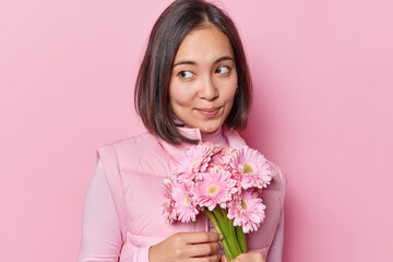 Portrait of pensive young Asian woman with thoughtful expression wears turtleneck and vest holds bouquet of gerbera flowers celebrates birthday isolated over pink background has romantic mood