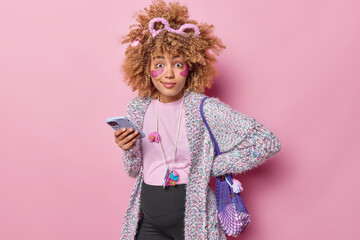 Photo of surprised curly pregnant woman poses with smartphone and toys in net bag surfs internet reads advice for future mothers applies hydrogel patches under eyes isolated over pink background