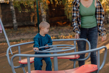 Cute Little boy with down syndrome in a funny hat walks in the playground with his mother, spinning...