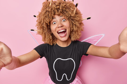 Happy positive surprised tooth fairy dressed in costume with wings keeps mouth widely opened arms outsreched makes photo of herself exclaims from joy isolated over pink background. Imaginary character