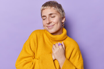 Young pleased woman keeps hands to heart as hears pleasant words or receives compliment stands with...