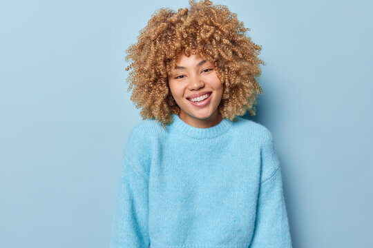 Portrait of happy curly haired woman smiles broadly shows white teeth dressed in long sleeved jumper looks directly at camera isolated over blue background. People and positive emotions concept