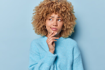 Fototapeta na wymiar Photo of thoughtful beautiful European woman with curly hair keeps hand on chin concentrated aside has pensive expression dressed in casual jumper isolated over blue background. Let me think