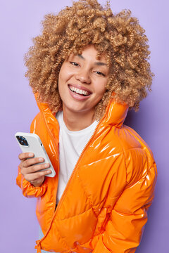 Happy curly teennage girl dressed in warm orange jacket smiles broadly holds cellphone checks notification going to have walk outdoors expresses positive emotions isolated over purple background