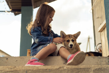 Side view of pretty little girl with long dark hair wearing denim jacket, caressing, stroking fur of welsh pembroke corgi puppy pet, sitting outside on concrete stairs near house in yard. Pet care.