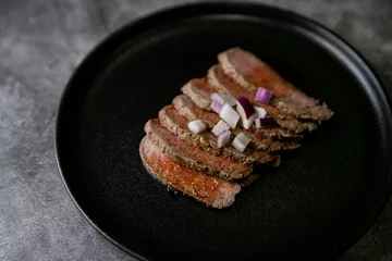 Foto op Canvas Closeup shot of steak tataki in sauce and onions on black plate with grey blurred background © Metanoiamoments/Wirestock Creators