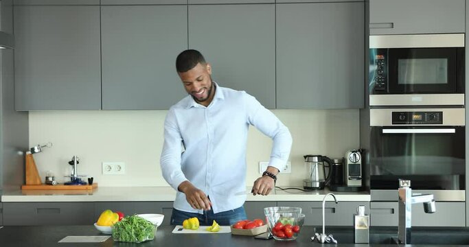 Happy cheerful handsome African cook blogger, chef guy dancing to music at organic food on kitchen table, slicing vegetables for salad, preparing ingredients, smiling, having fun, enjoying activity,
