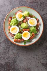 Homemade salad of steamed romanesco cabbage, boiled eggs, fried bacon and fresh microgreens close-up in a plate on the table. Vertical top view from above