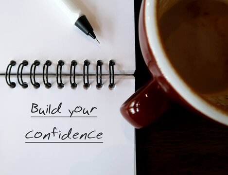 Coffee cup and notebook with handwritten text BUILD YOUR CONFIDENCE - to boost feeling of self-assurance, appreciation of self abilities which make sure of choices and decisions in life