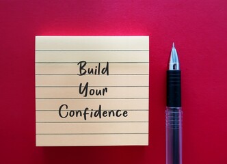 Yellow note on red background with handwritten text BUILD YOUR CONFIDENCE - to boost feeling of...