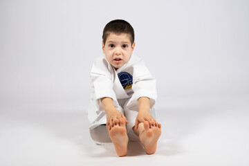 A little boy in a kimono doing karate, stretches his hands to the tips of his toes, practices...