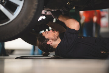 Fototapeta na wymiar Young Asian male wearing uniform and hand gloves while in modern garage inspecting and working on maintenance and repair of car by lying on floor under the car and checking underneath the vehicle
