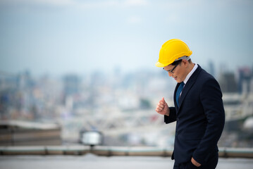 Young business man and architect or contractor in formal clothing with yellow hardhat standing with open arms wearing eyeglasses on rooftop of new building against tall buildings