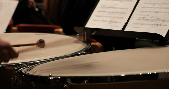 Close up shot of a drummer with a timpani during a concert with orchestra to classical music for the mozart requiem while the drumsticks are beating with sheet music in the background. Slow motion