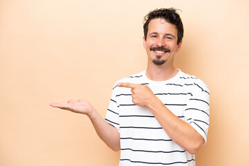 Young caucasian man isolated on beige background holding copyspace imaginary on the palm to insert...