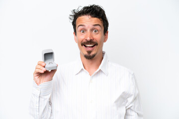 Young man holding a engagement ring isolated on white background with surprise and shocked facial...
