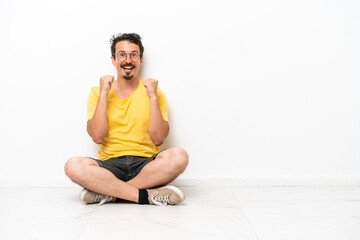 Fototapeta premium Young caucasian man sitting on the floor isolated on white background celebrating a victory in winner position