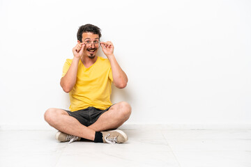 Young caucasian man sitting on the floor isolated on white background with glasses and surprised