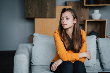 Disappointed brunette hispanic woman in orange blouse sitting on cozy sofa with upset facial expression, tired frustrated girl after divorce failure mistake. Perplexed lady sitting on sofa at home.