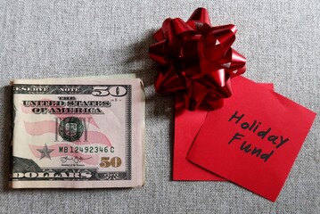 A red gift bow on red sticky note with text written HOLIDAY FUND on cash dollars money on wood background , concept of Christmas New Year Holiday season gifts shopping budget