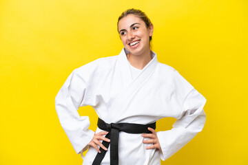 Young caucasian woman doing karate isolated on yellow background posing with arms at hip and smiling