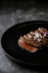 Keuken spatwand met foto Closeup shot of steak tataki in sauce and onions on black plate with grey blurred background © Metanoiamoments/Wirestock Creators