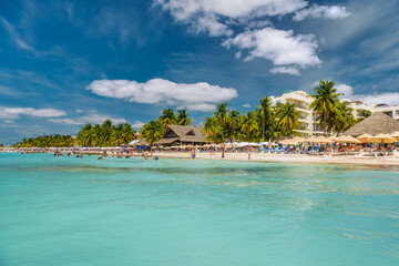 People swimming near white sand beach with umbrellas, bungalow bar and cocos palms, turquoise...