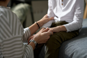 Close-up of young mother supporting her little son holding his hands while they sitting and talking in the room
