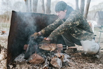 young man survivalist cooks roasts chicken meat food are fried on grill on smoldering coals or ember from a campfire on the ground. barbecue in camping conditions. countryside and wild rustic life