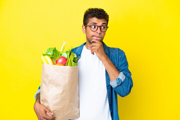 Young Brazilian man holding a grocery shopping bag isolated on yellow background having doubts and...