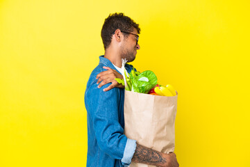 Young Brazilian man holding a grocery shopping bag isolated on yellow background suffering from...