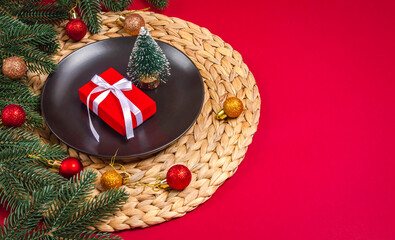 Christmas gift on a black plate, green fir branches with Christmas balls on a red background with space for text.