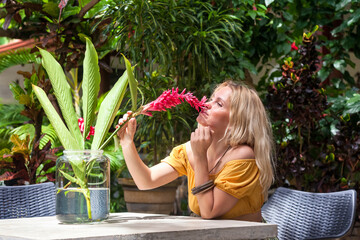 Happy relaxed attractive blonde woman in trendy summer wear, sniffs smell flower tropical red...