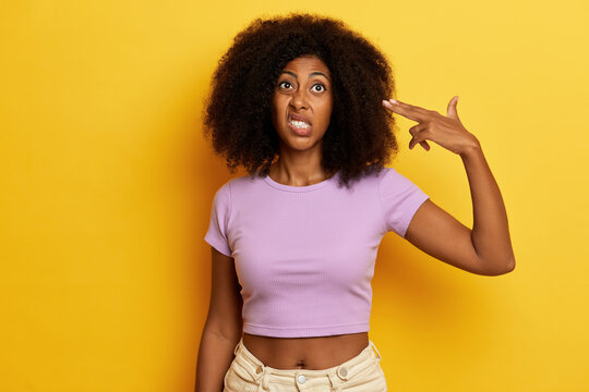 Photo of disappointed curly haired woman in purple T-shirt, makes finger gun pistol close to her temple, isolated on yellow background