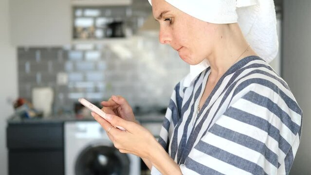 portrait of a woman with a towel on her head looking in the phone standing in the kitchen