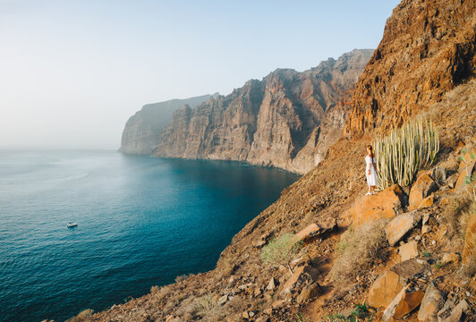 Landscape photo of a woman standing at the edge of big cliffs, the beach and the ocean in the background. Los Gigantes. Incredibly beautiful turquoise water. Tenerife, Canary island, Spain