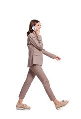 Happy young woman in formal suit talking on smartphone while walking against white background