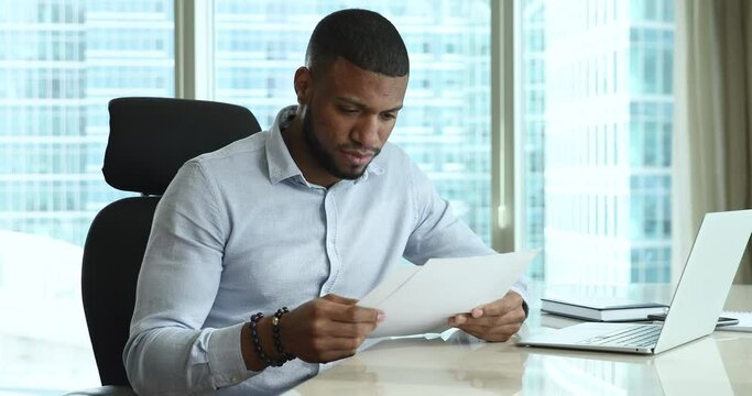 Serious Focused African Employee Man Holding Document, Receiving Letter, Reading Legal Paper, Reviewing Project Report, Sitting At Laptop In Office With Panoramic Window. Paperwork, Business Concept